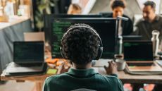 Female software developer working on a desktop computer with fellow software engineers collaborating in background.