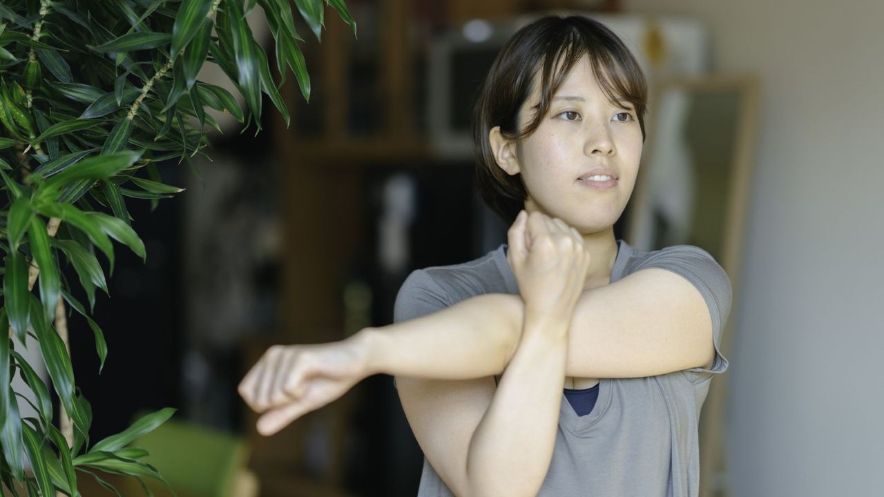 A woman in a grey t-shirt stretches her shoulder with an arm across her body 