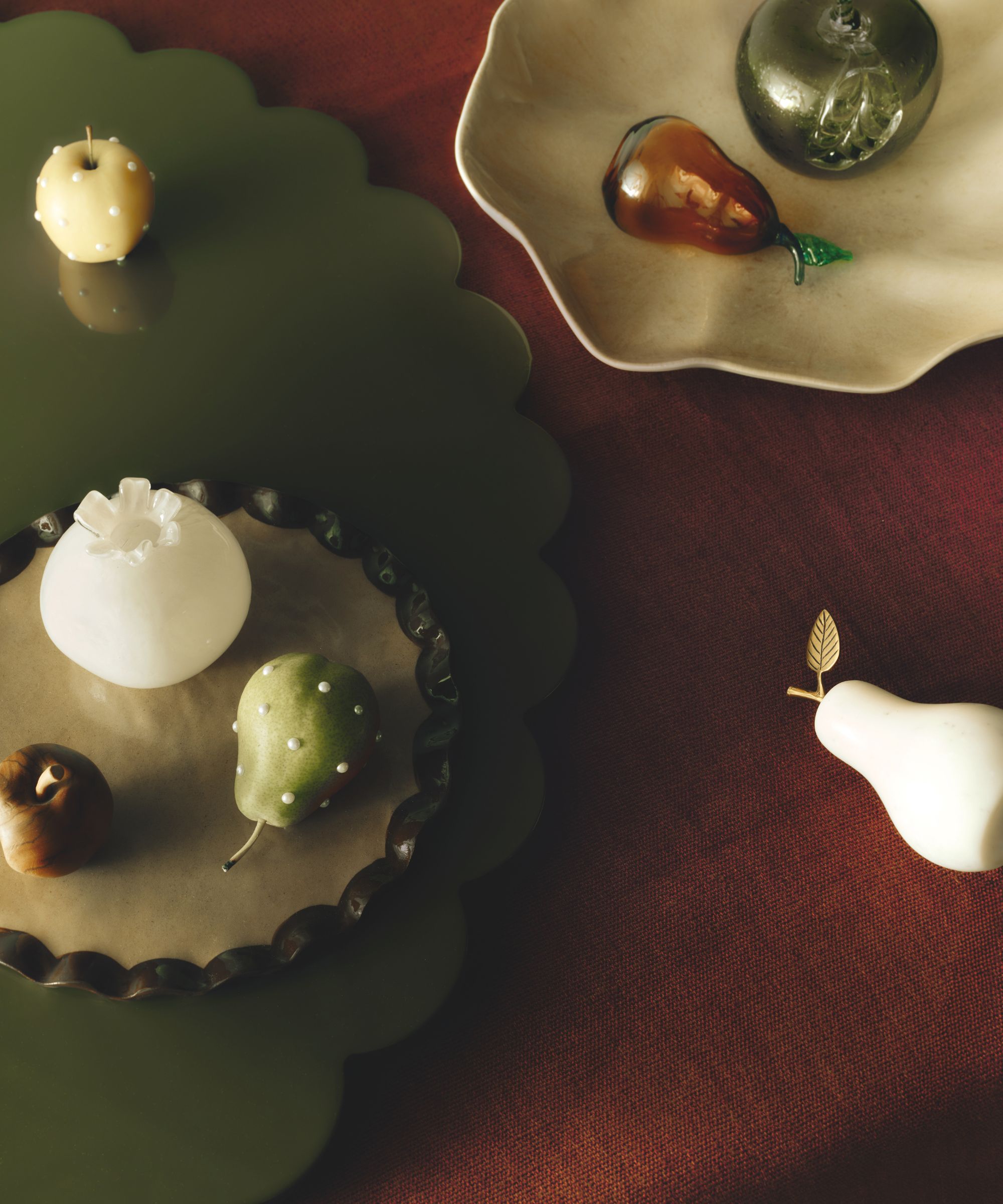 An overhead shot of decorative glass and stone fruits, including a pear with pearl accents and a marble pear with a gold leaf, resting on scalloped green and white plates.