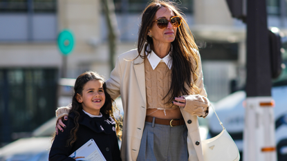 Chloe Harrouche wears sunglasses, a white ripped shirt from Miu Miu, a brown braided wool pullover from Miu Miu, a beige long coat, a brown shiny leather belt from Miu Miu, gray large wide legs pants, white leather ripped sneakers, and is seen with her daughter, outside Miu Miu , during Paris Fashion Week - Womenswear F/W 2022-2023, on March 08, 2022 in Paris, France.