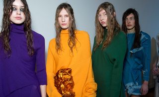 Row of young female models, rainbow hue clothing, long straggly hair style, eye makeup, white wall