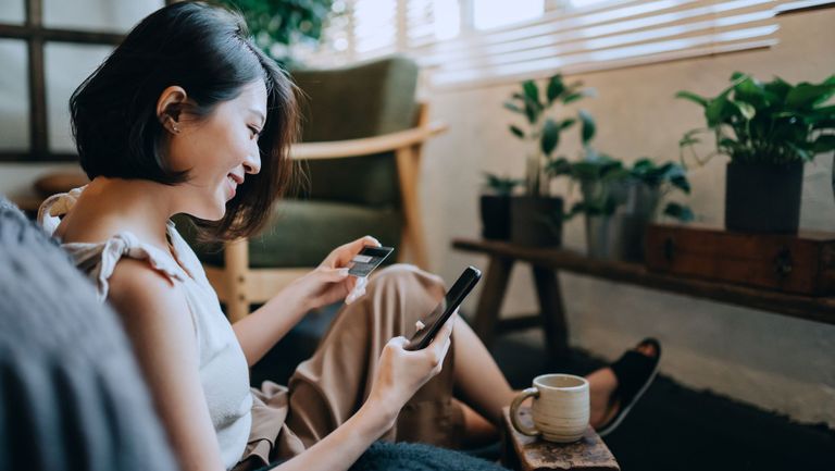 beautiful smiling young asian woman chilling at home, sitting on the floor in bedroom, enjoying a cup of coffee and shopping online with smartphone while making mobile payment with credit card on hand