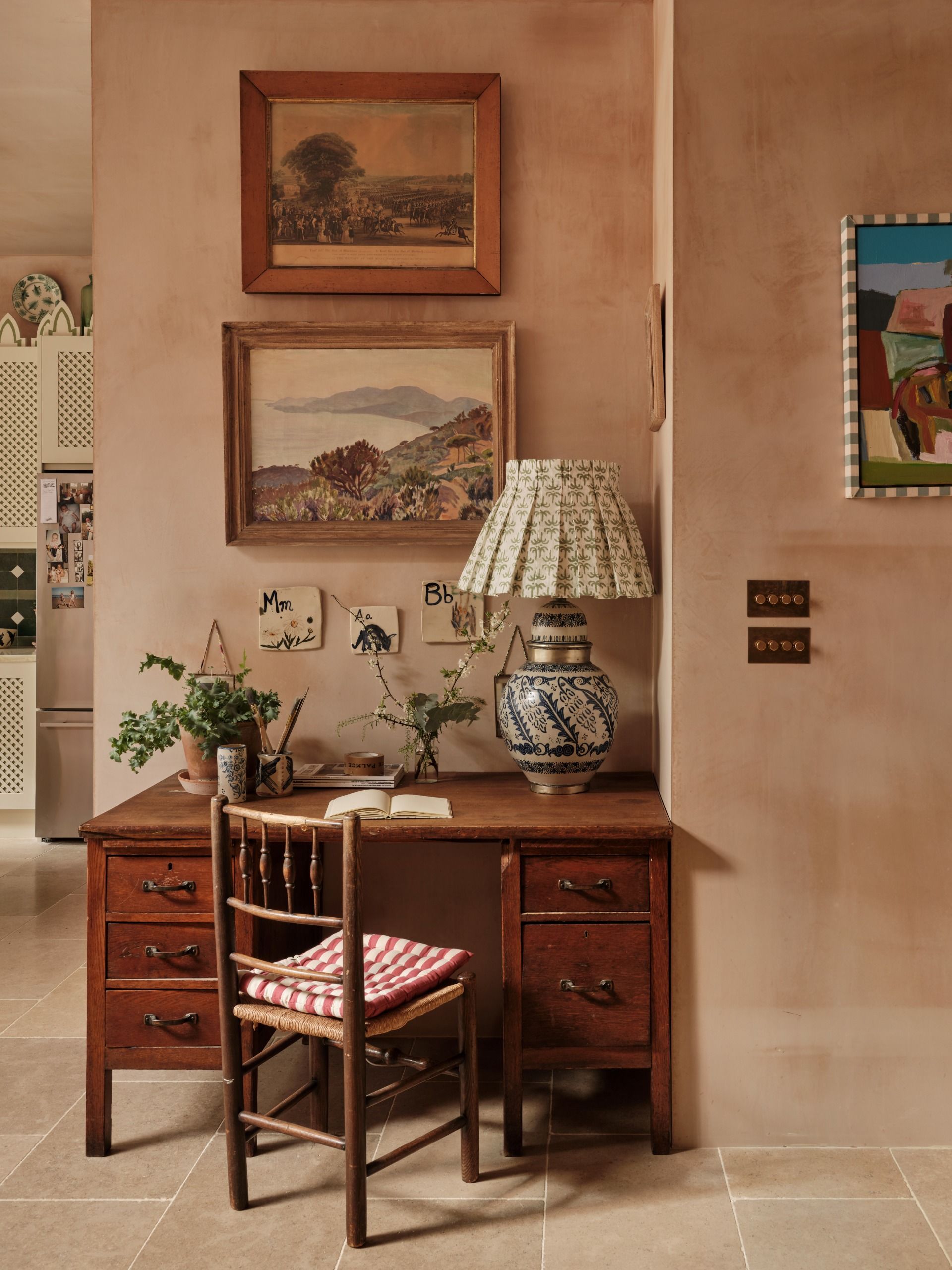 A home nook decorated with vintage furniture like a wooden desk with organizer drawers, a vintage lamp, potted plants, and artworks on argilla walls.