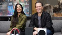 Xbox CEO Asha Sharma sat on a sofa next to ex-CEO Phil Spencer, both smiling with a variety of Xbox-themed objects in the room, including consoles on the desk and a poster in the background.