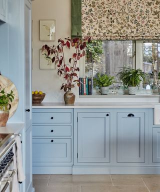 Pale blue kitchen with painted cabinets, floral green blind, white countertops, stone floor tiles, plants, black hardware, artwork