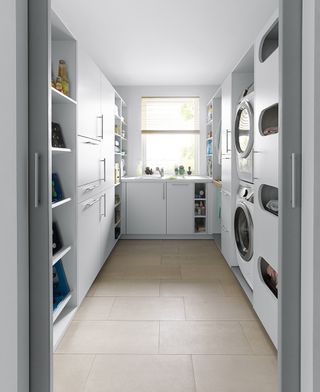 all-white utility room by Schüller with a sink by the window, built-in washing machine and tumble dryer, and open shelving units