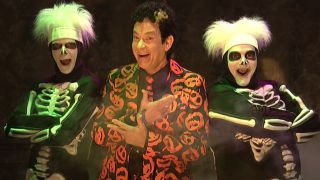 Tom Hanks as David Pumpkins smiling and pointing at his two dancers dressed as skeletons.