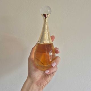 A close-up of Digital Beauty Writer, Naomi Jamieson's hand holding Dior's J'adore L'or essence de parfum, in front of a cream wall