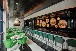 The interiors of a restaurant feature space-age design furniture in eye-catching tones of green mixed with white and black tiles flooring, a sleek bar counter featuring spherical drink displays, and 1970s-lighting.