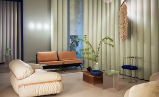 Interior view of the lounge at Studiopepe’s Club Unseen. The space features peach and light pink seating, a blue and silver chair, a low table with a plant in a green vase and other items, a round light green side table and a pink fluffy wall hanging suspended in front of a pastel green tubular wall