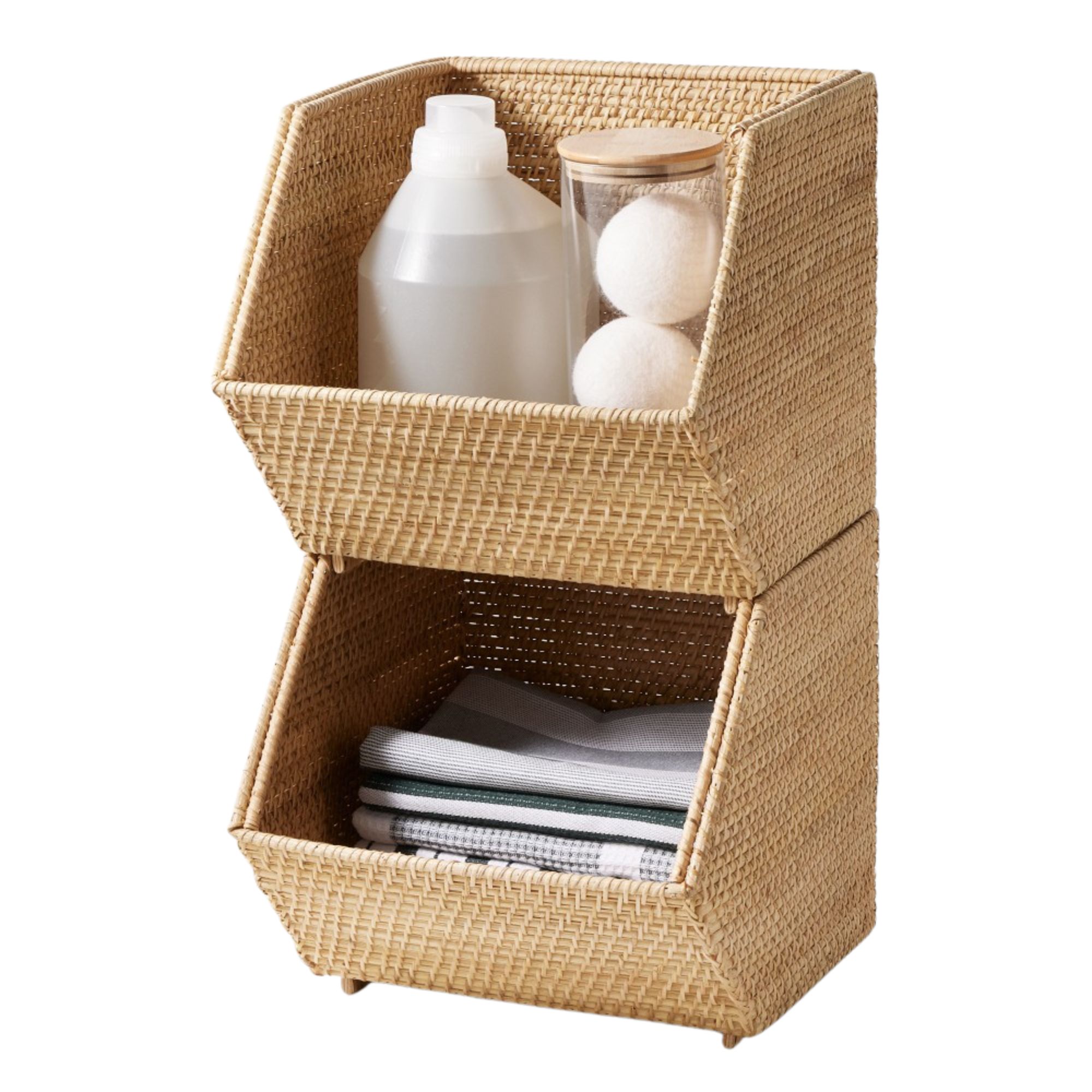 Two stacked rattan organizers. The lower one has folded hand towels, and the top one has an unmarked bottle of liquid detergent, and a glass jar with wool air dryer balls