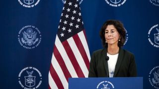 US Secretary of Commerce Gina Raimondo answering questions at a news conference, stood at a podium. Behind her, the US flag hangs from a pole and a backdrop carries the repeated emblem of the US Department of Commerce.