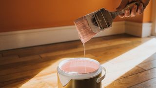 Hand dipping a paint brush into a large tin of pink paint as it drips back into the tin. Paint is one source of volatile organic compounds (VOCs) in your home.