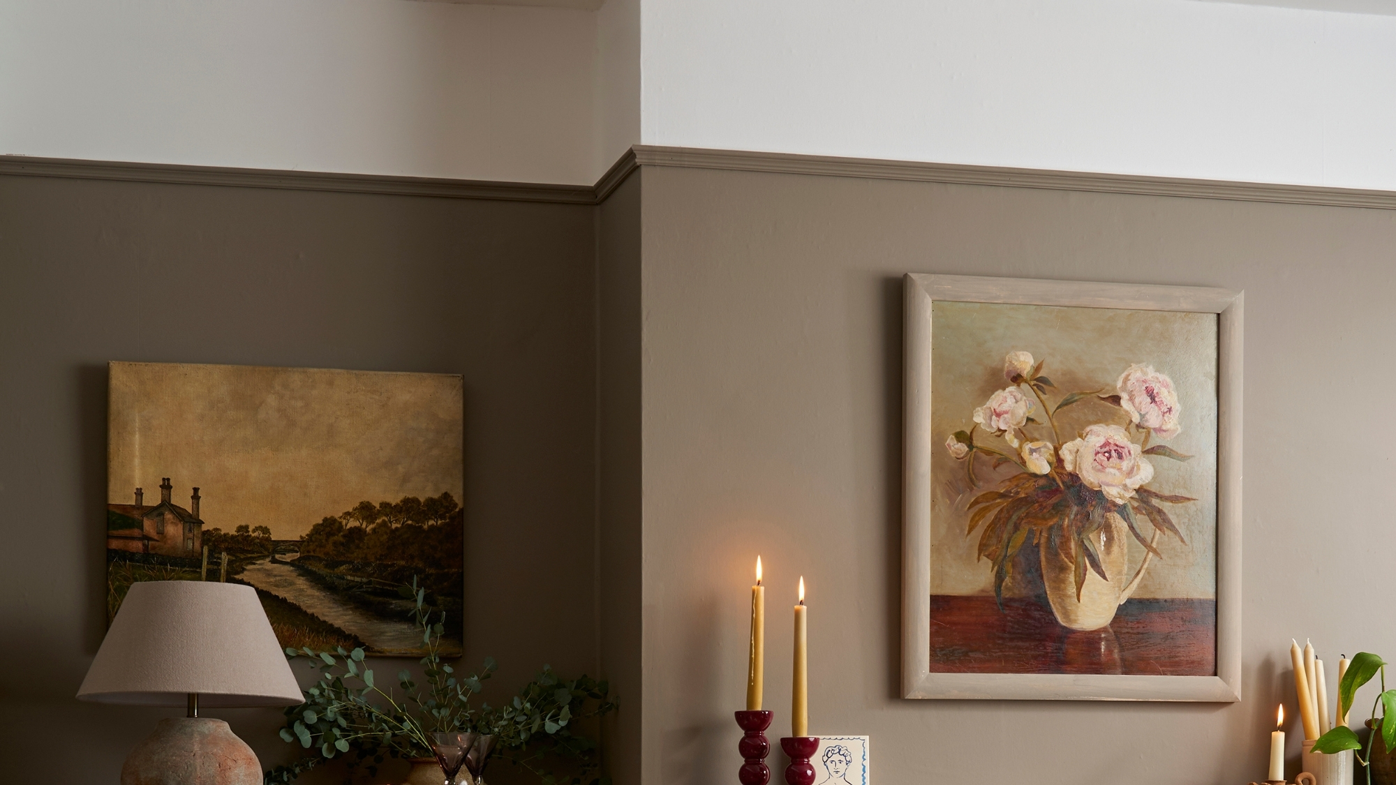 Two-tone brown walls with a picture rail, floral and landscape paintings, candles, and a lamp.