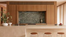 A beautiful kitchen with wood cabinets, a green stone backsplash, a limewash island, three stools, and an ikebana arrangement on the counter by the sink