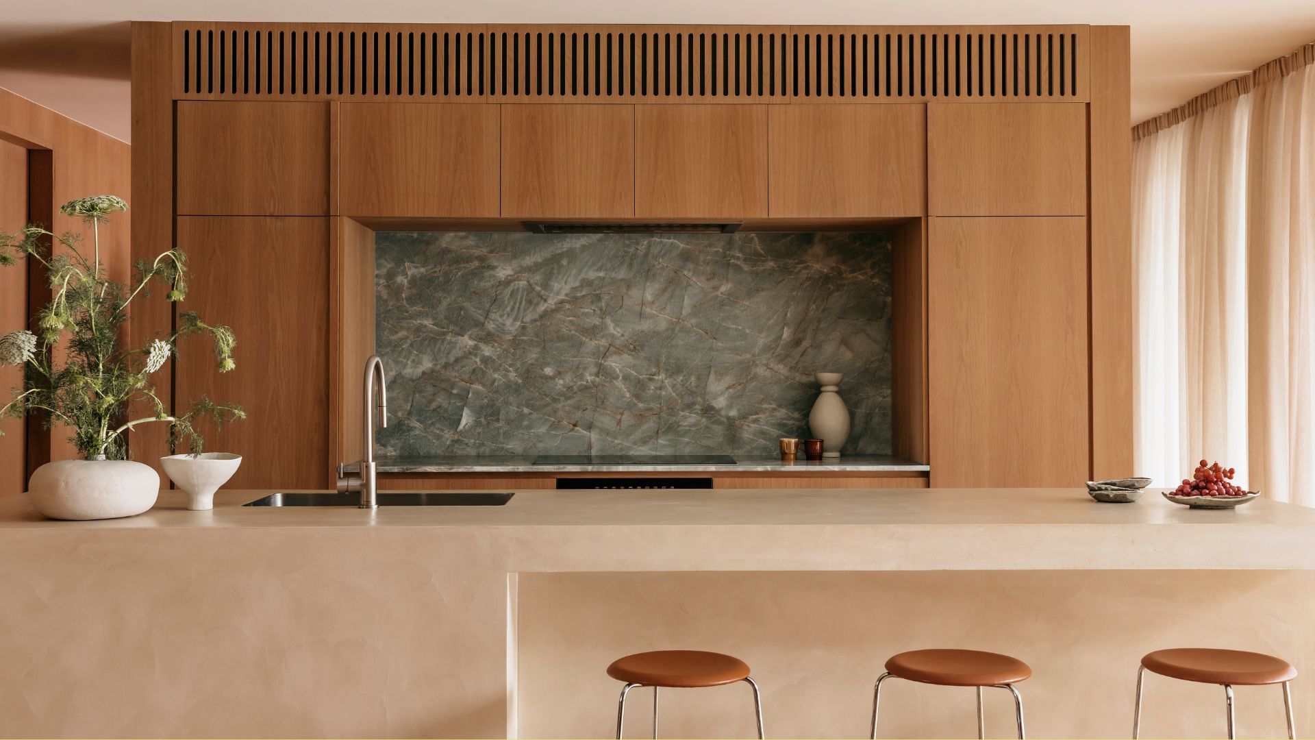 A beautiful kitchen with wood cabinets, a green stone backsplash, a limewash island, three stools, and an ikebana arrangement on the counter by the sink