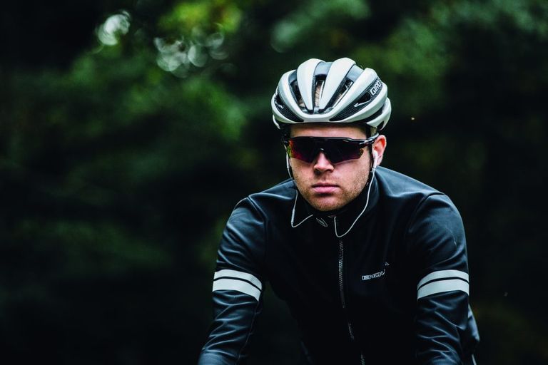 Should you cycle with earphones in? This image show's a rider heading towards a camera with a white helmet on, earphones with wires, dark glasses and a black rain jacket on 