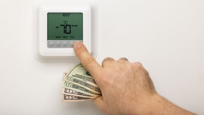A hand holding money adjusting a thermostat on a wall.
