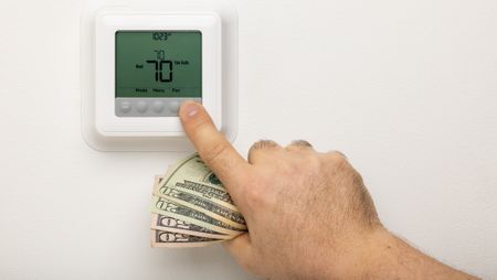 A hand holding money adjusting a thermostat on a wall.