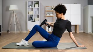 Woman using massage gun, she is sitting on an exercise mat