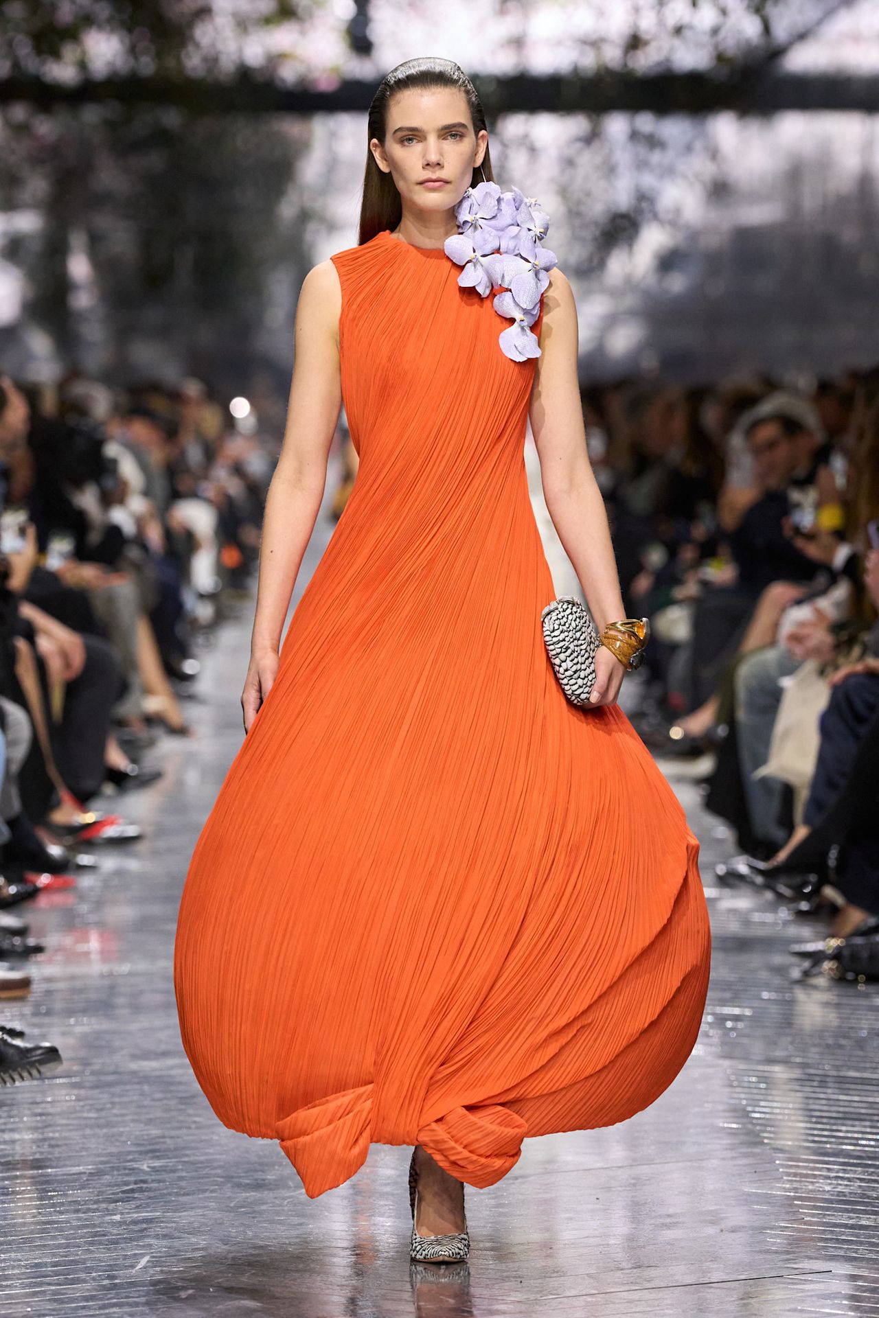 A Christian Dior Haute Couture model wearing a voluminous pleated burnt tangerine gown adorned with purple orchids styled with a textured leather gray clutch bag and a pair of textured leather pointed toe gray pumps.