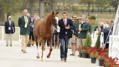 Boyd Martin, a rider wearing a blue blazer, red tie and jeans, runs beside his chestnut horse Neville Bardos, at the horse inspection at the 2010 World Equestrian Games
