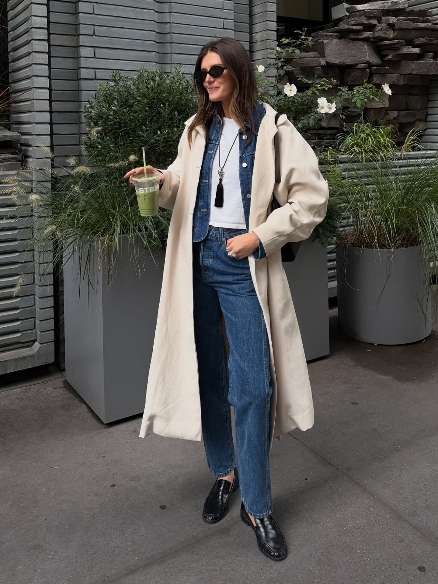 NYC style influencer Coco Schiffer poses with a green juice wearing a trench coat, denim jackets, white T-shirt, long pendant necklace, straight-leg jeans, and black ruched loafers.