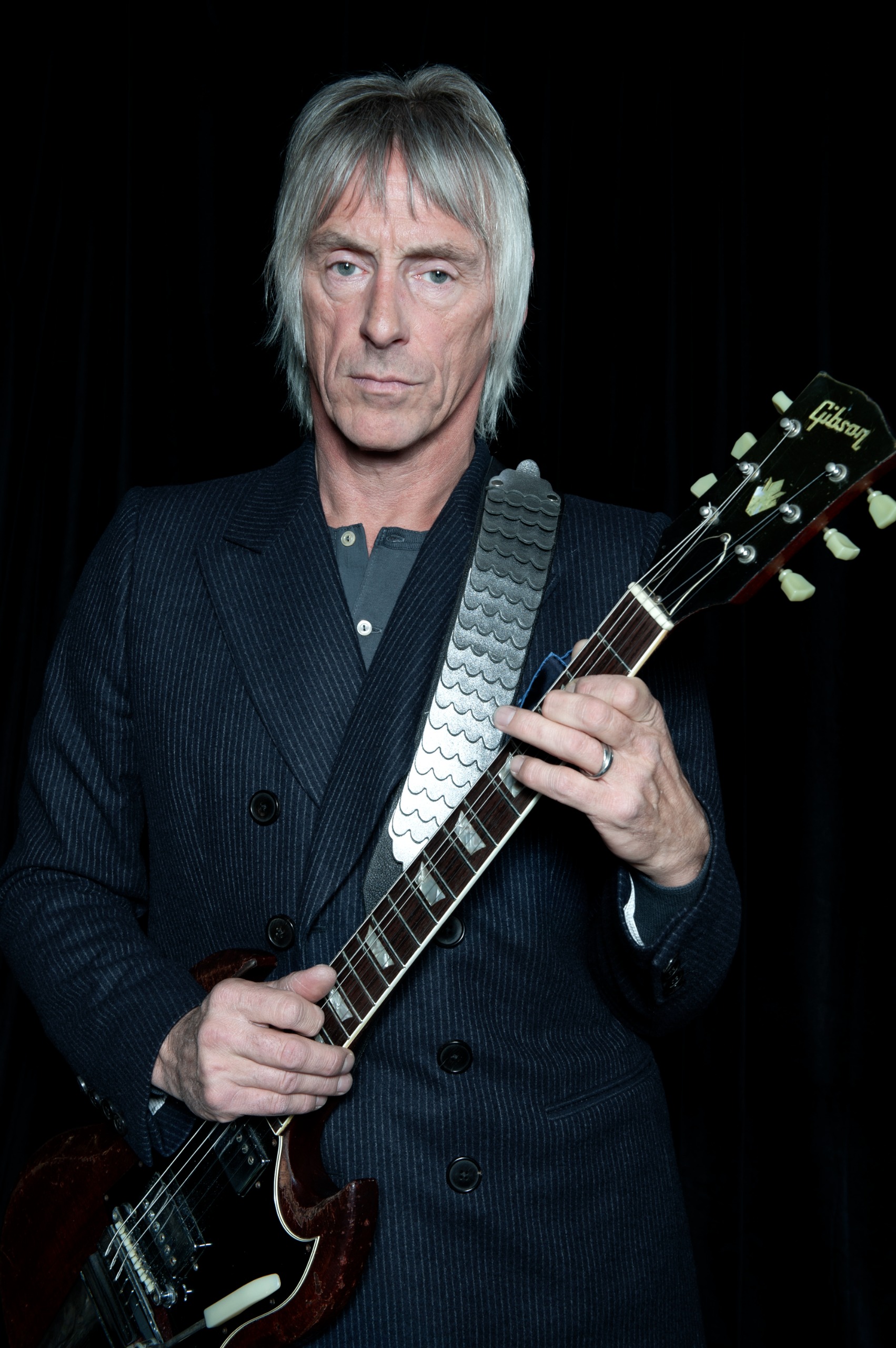 Paul Weller, in a pinstripe suit, holds a well-worn Gibson guitar close to the camera in a moody studio portrait against a black background.