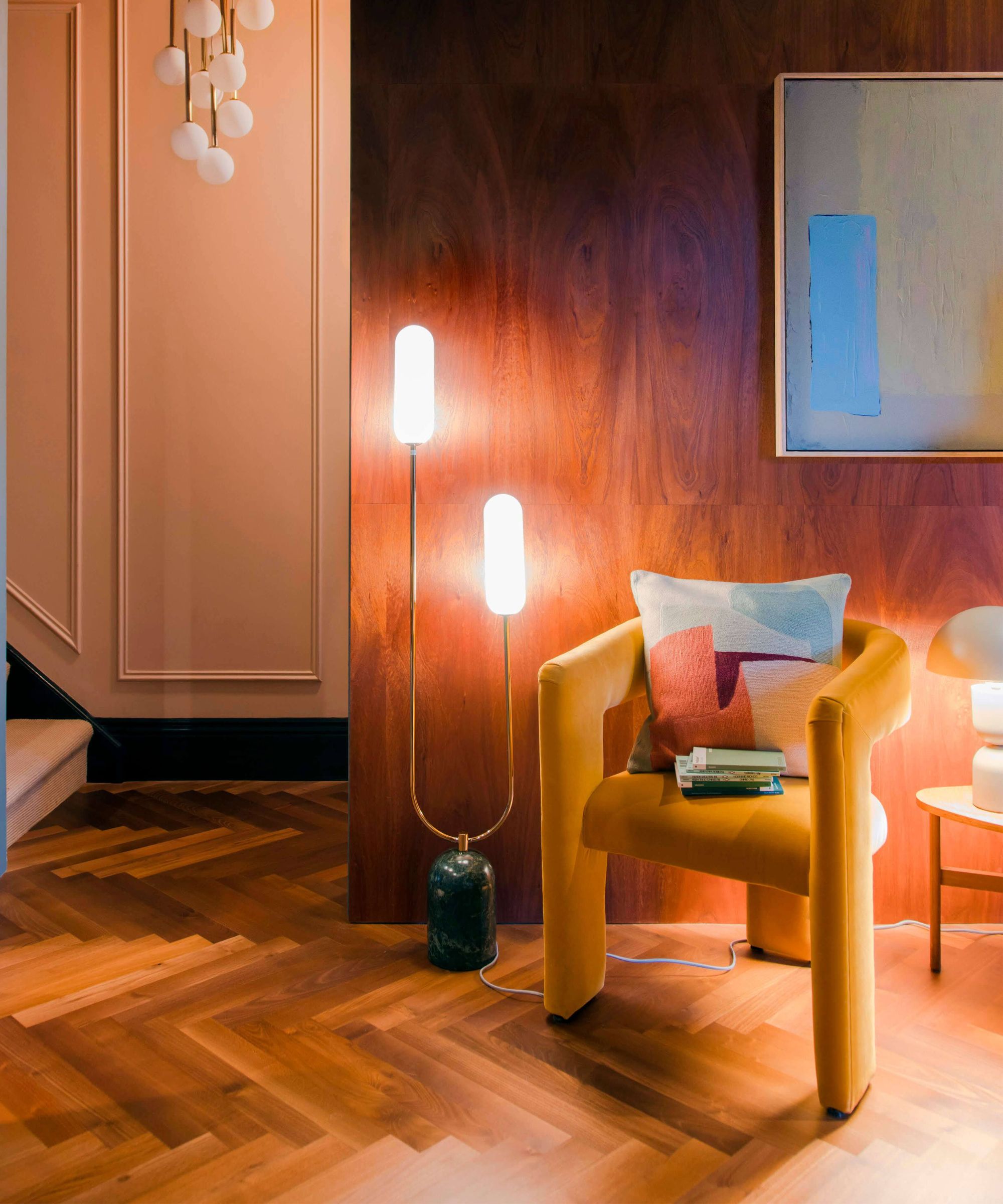 A cosy corner with a mustard-yellow armchair topped with a patterned cushion, placed beside a glowing twin-bulb floor lamp with a marble base. The setting features warm wood panelling, parquet flooring, and soft ambient lighting, creating a welcoming reading nook.