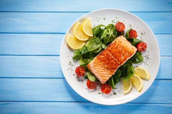 a filet of salmon sits on an array of leafy greens and half-cut cherry tomatoes, crowned on opposite sides by thin quarter lemon slices, all on a white plate that sits on a blue wood table.