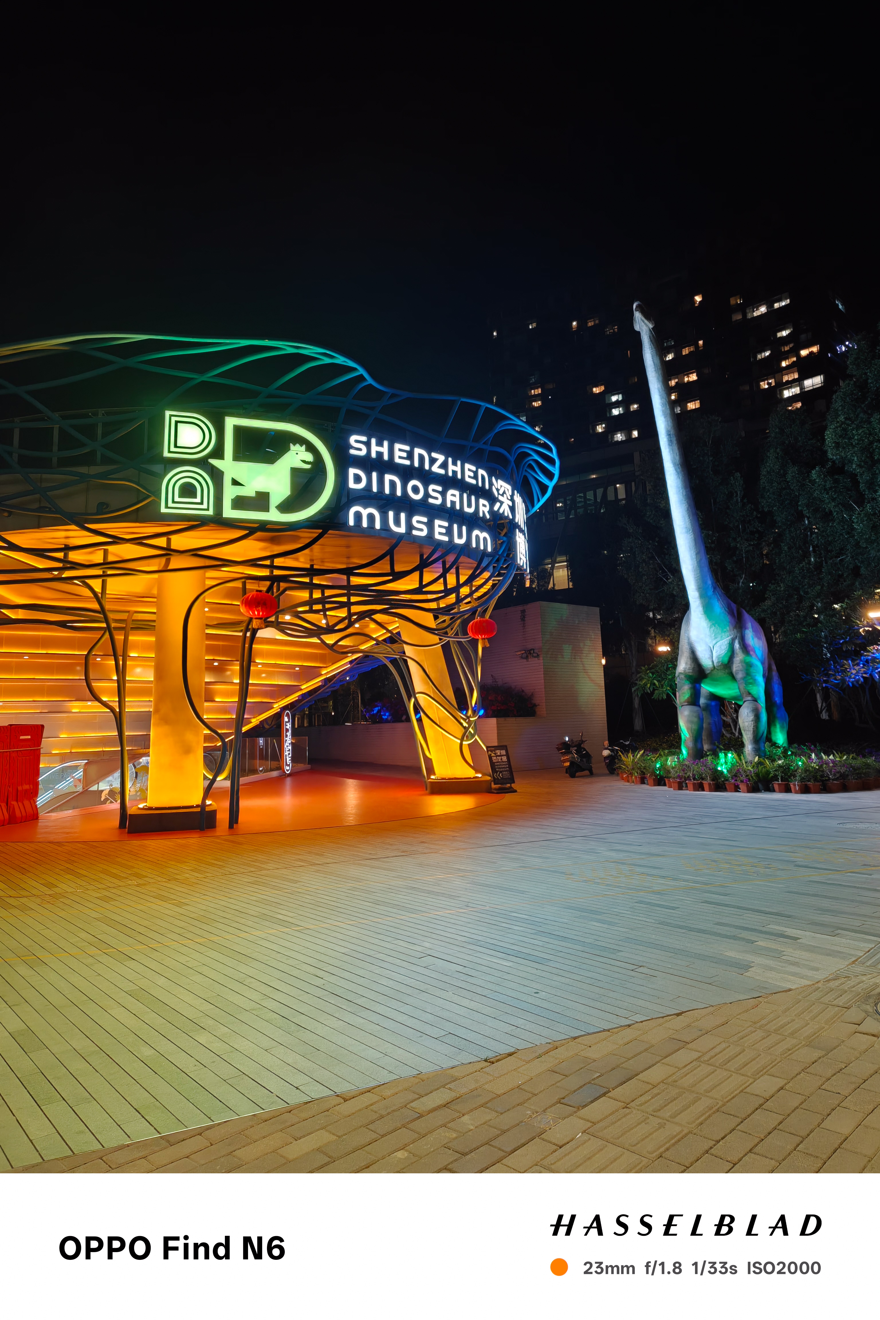 A wide night shot of the entrance to the "Shenzhen Dinosaur Museum." The entrance features a glowing yellow organic structure with the museum's logo in green neon. To the right, a large, life-sized Brachiosaurus statue stands tall under the city lights.