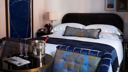 A shot of a double bed in a room in the hotel NoMad London. Sparkling wine sits in an ice bucket on a side table in the forground, and in the background is one of NoMad's signature double beds