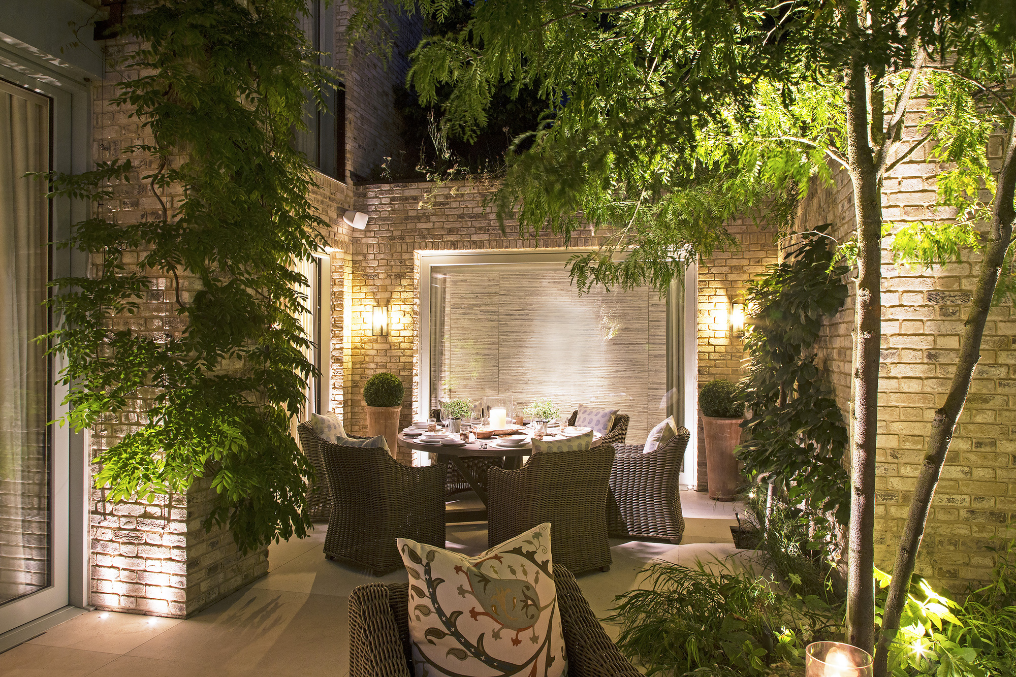 A patio area with a circular dining table, wall mounted lanterns on a brick wall, and trees with uplighting
