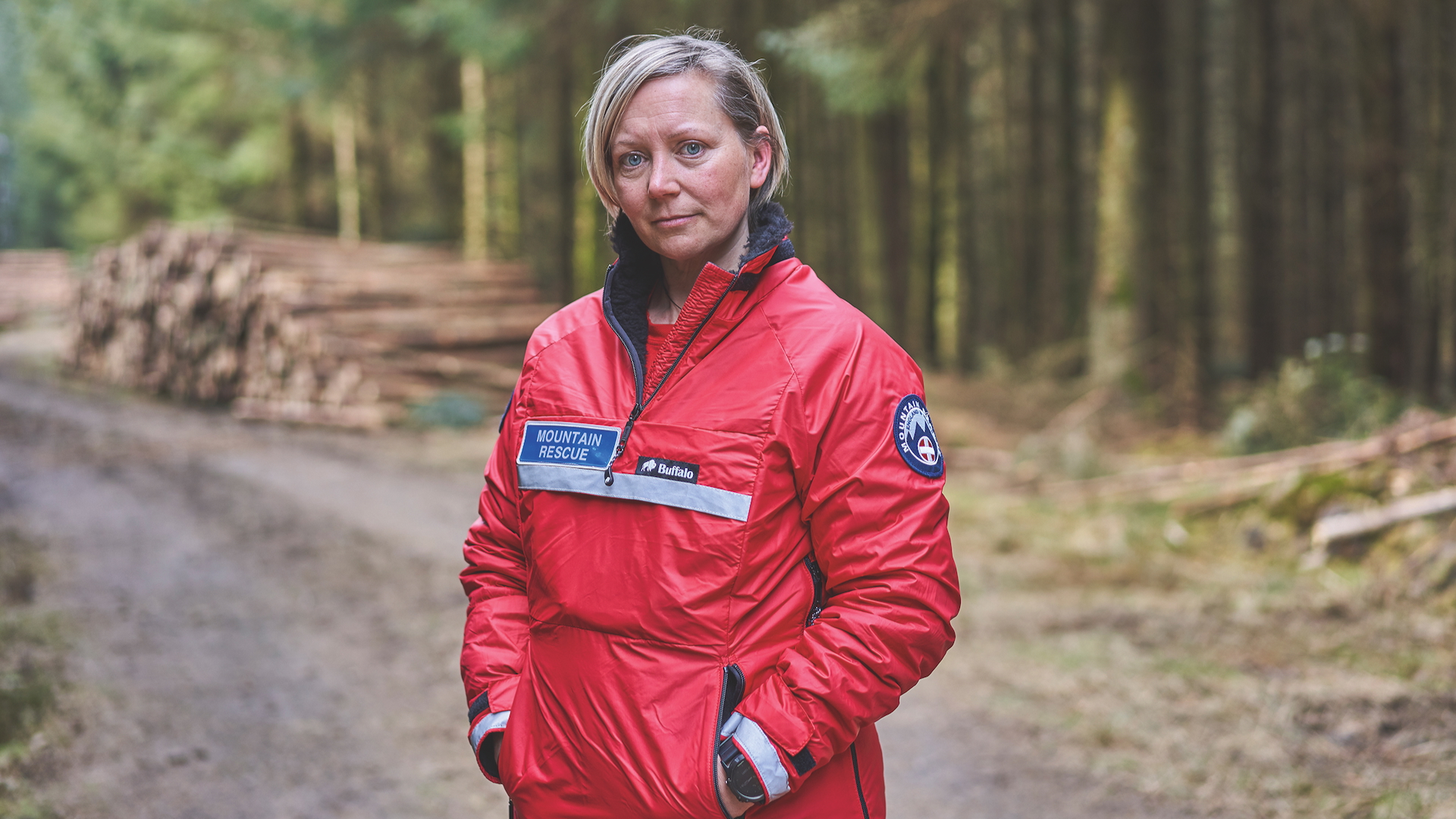 Mel Smith from Huddersfield stands in a forest wearing her uniform for Holme Valley Mountain Rescue Volunteer Team, Yorkshire