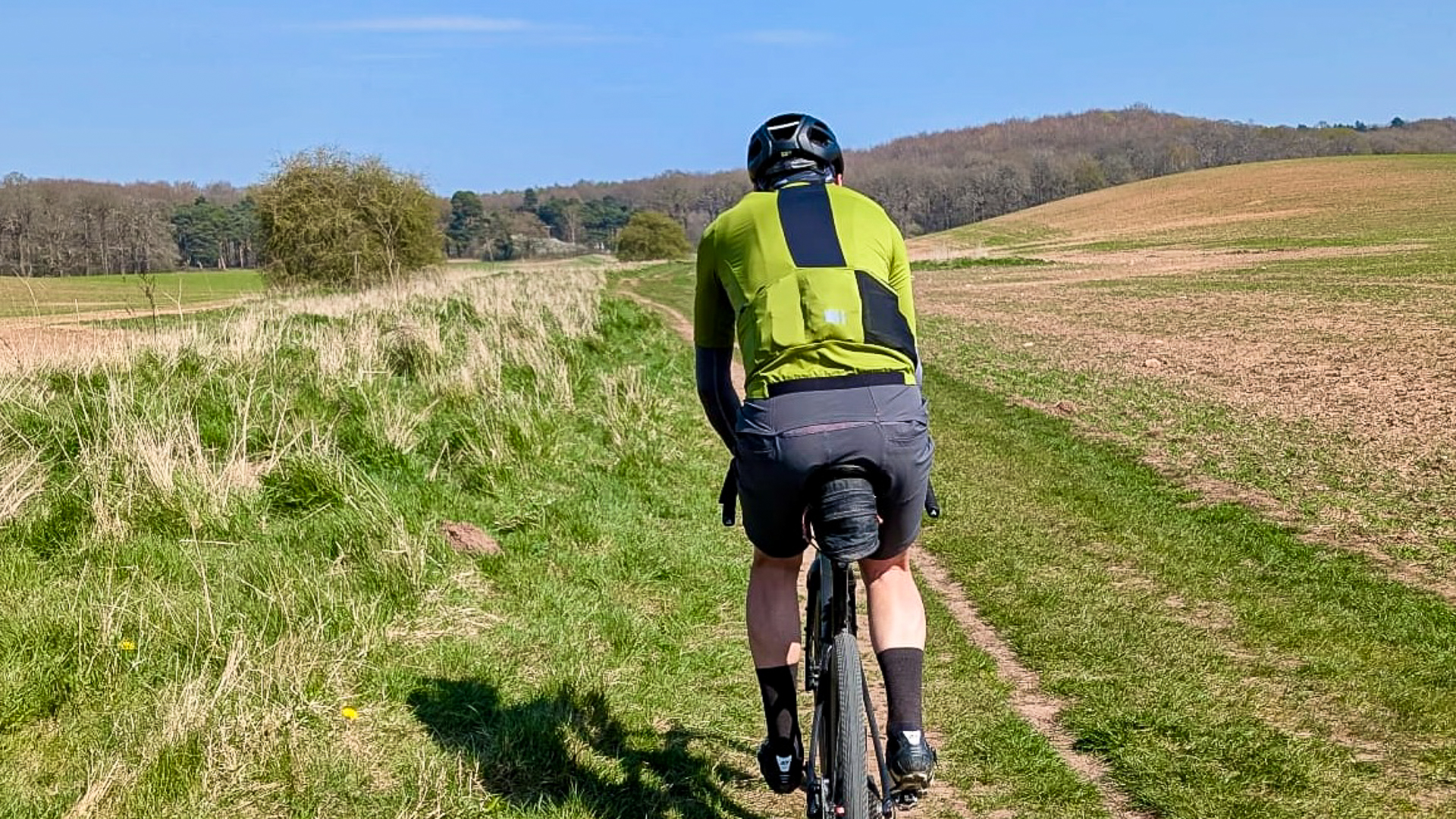 Rear shot of a man wearing a green jersey and grey shorts riding along a trail through a grassy field