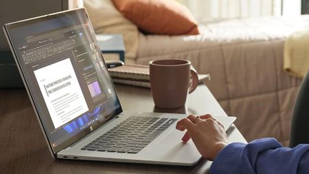 A person edits a document on a laptop beside a coffee mug in a sunlit living room, creating a calm, focused work-from-home scene.