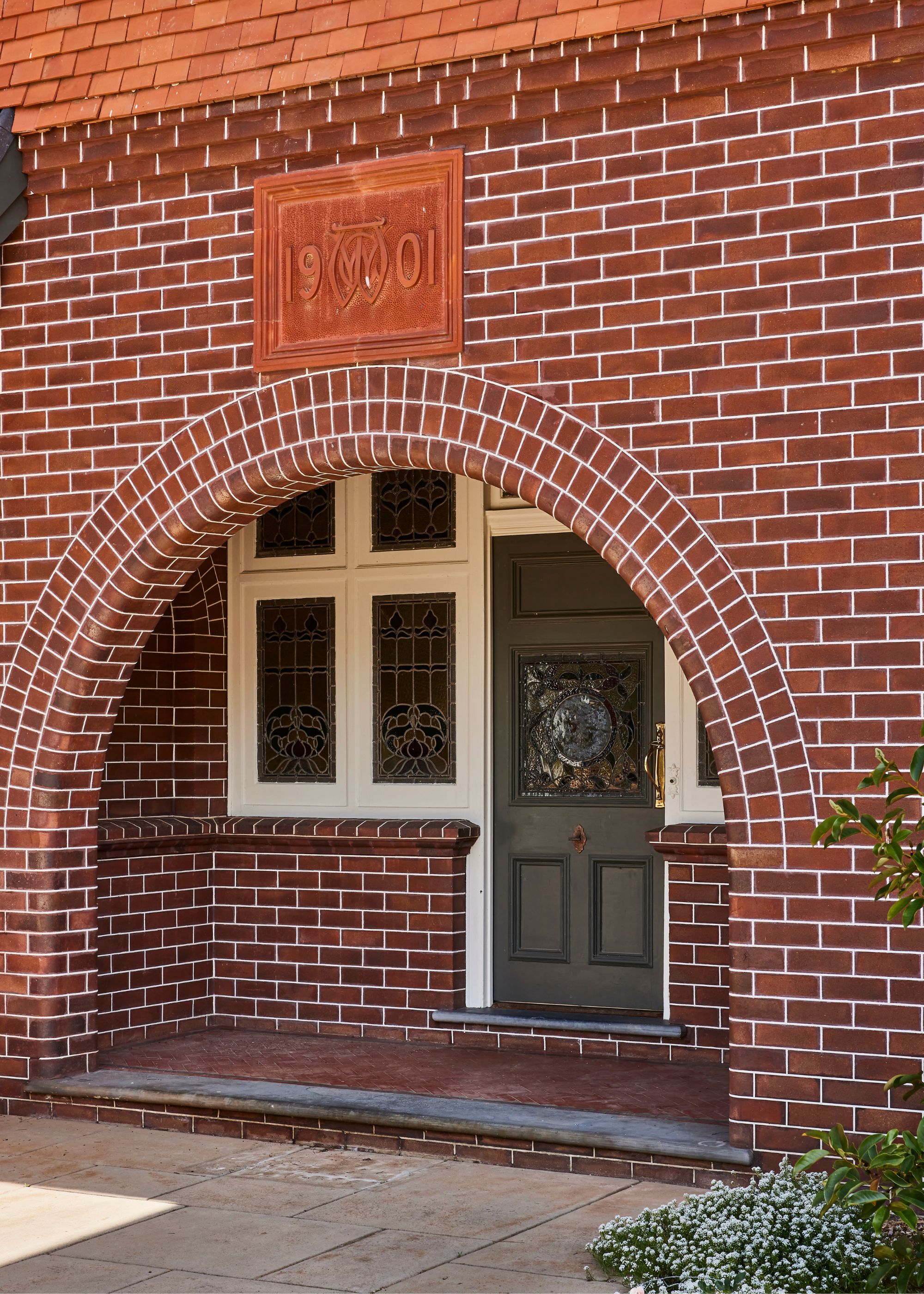 The exterior of a bricked home with a period style/traditional deep green English style front door as well as white framed windows.