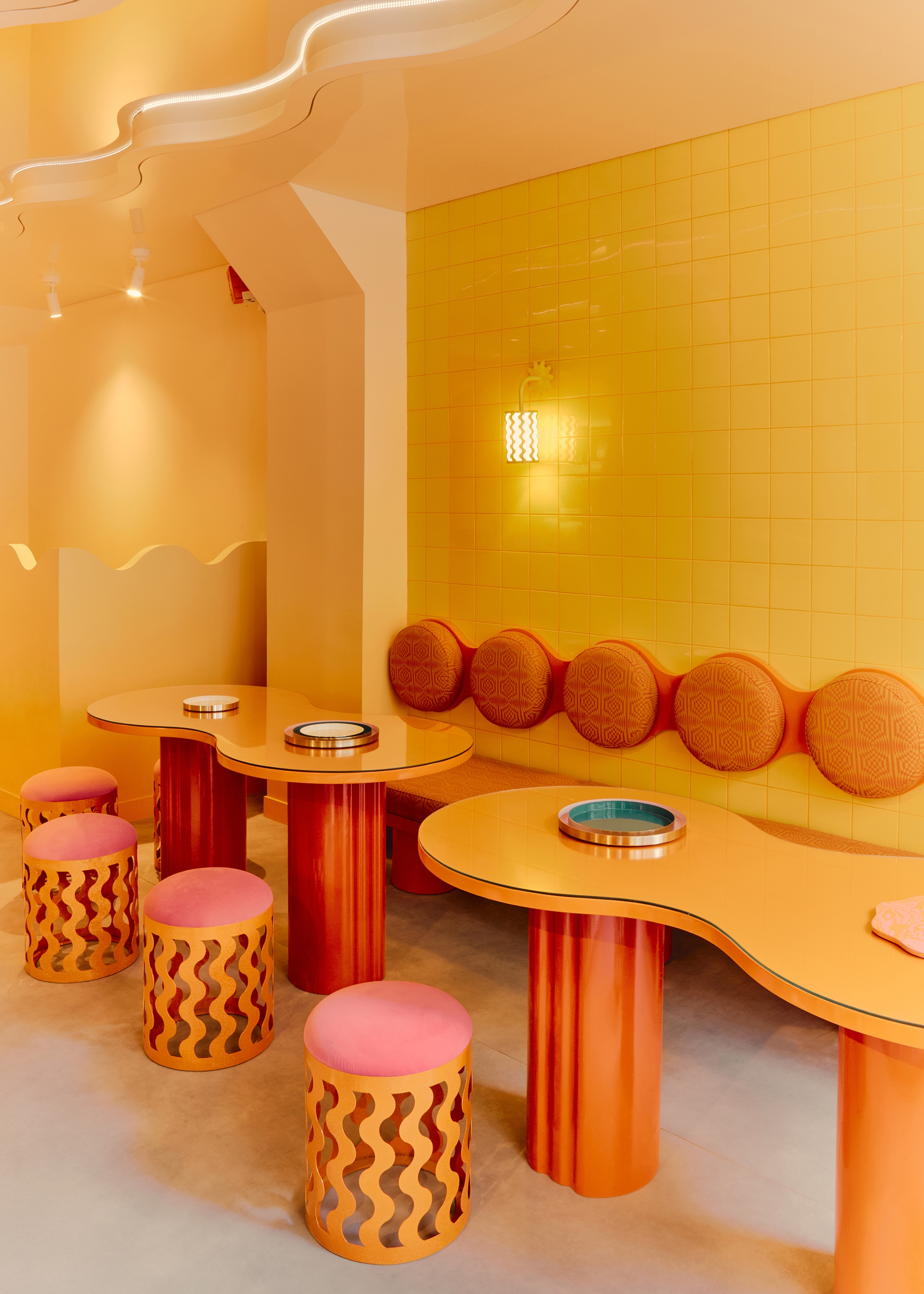 an immersive space with a warm orange and yellow color scheme with circle backrests along the banquette seating in front of circular tables joined at the centre with a wiggle format and circular stools with wavy bases as well as waved ceiling with lighting