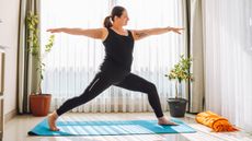A woman performs a yoga pose on a mat at home. She is in a lunge position, with her left foot forward and right foot back, left leg bent and right leg straight. Her arms are extended at shoulder height and she is facing her front left foot.