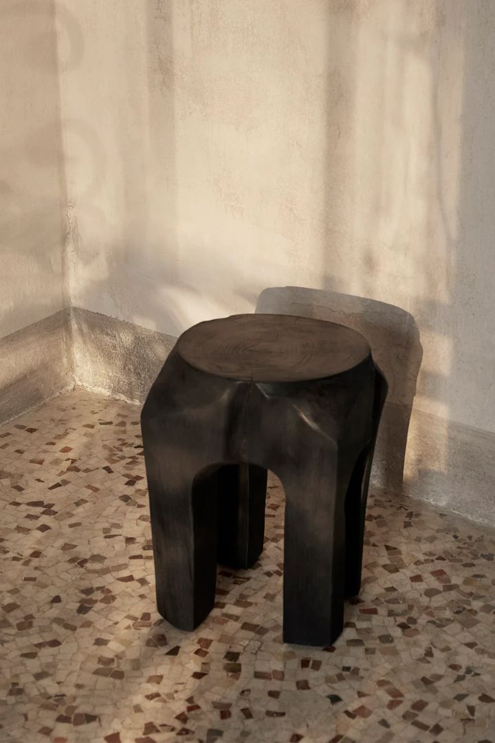 Ferm Living Root Stool in Black Stained, set atop terrazzo flooring in brown, beige, and black tile fragments in a sun-lit bare room.