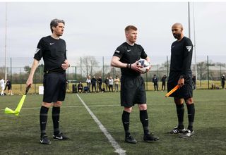Three match officials in black uniforms stand on a football pitch, preparing to officiate an under-19s fixture at the University of Warwick.
