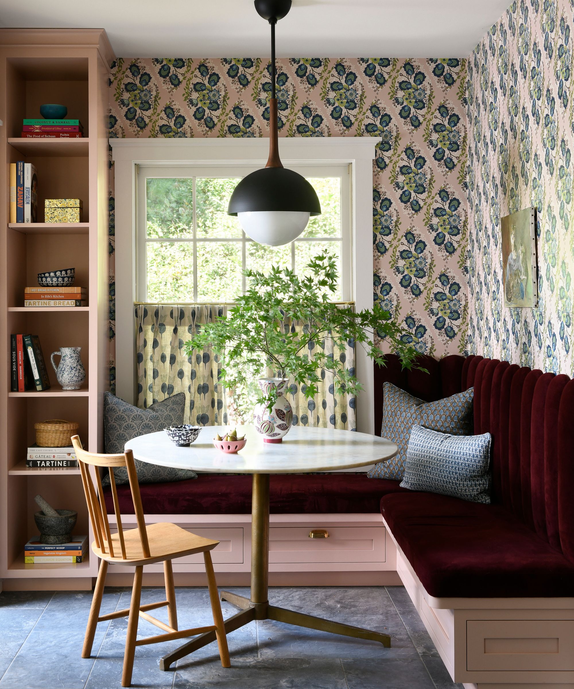A breakfast nook with dark red banquette seating, pink patterned wallpaper, and a window with patterned cafe curtains.