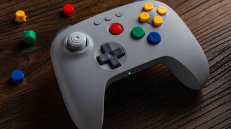 8Bitdo N64 controller in grey sitting on woodgrain desk next to buttons.