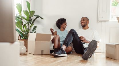 A smiling couple sit on the floor of their new home surrounded by moving boxes.