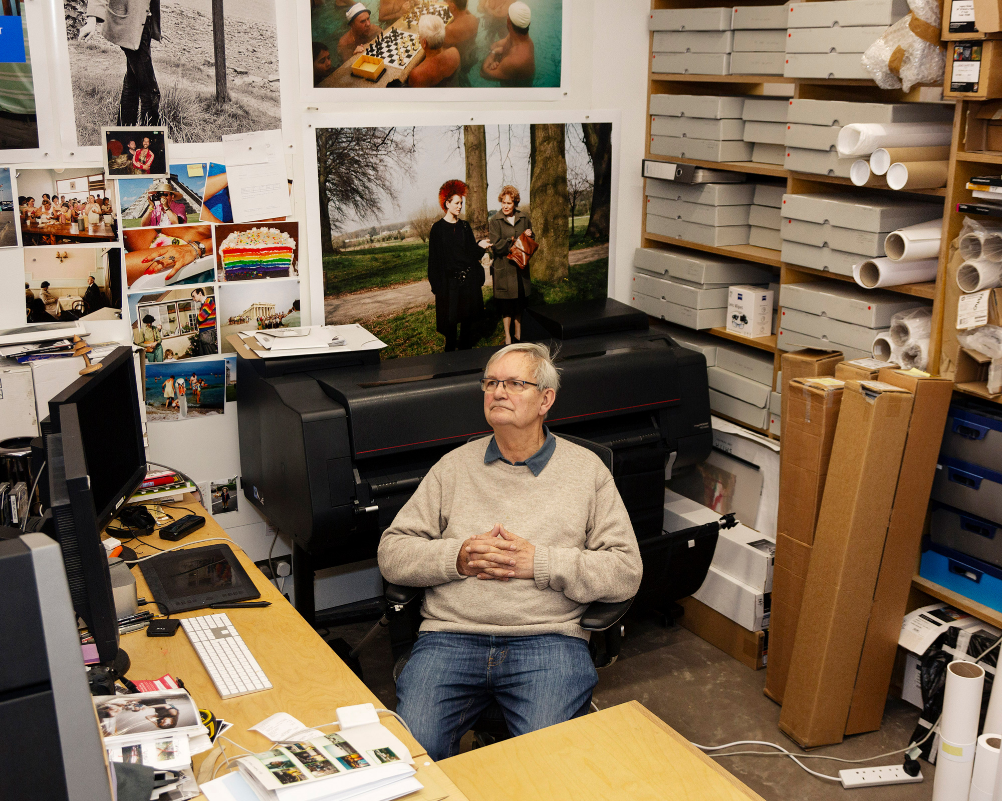 A middle-aged Martin Parr sits at a cluttered desk in an office or studio, looking upwards, surrounded by large printers, boxes, and photographs on the walls.