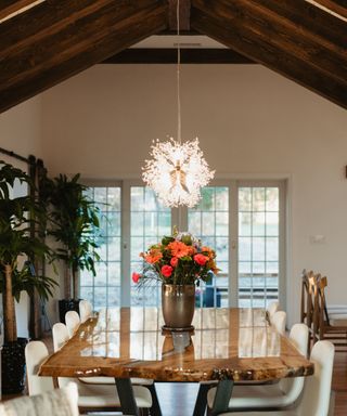 A farmhouse dining room with dark wooden beams on the ceiling, a white chandelier, a glossy wooden table with a vase of orange roses on it, with white leather chairs underneath it and tall green plants to the left
