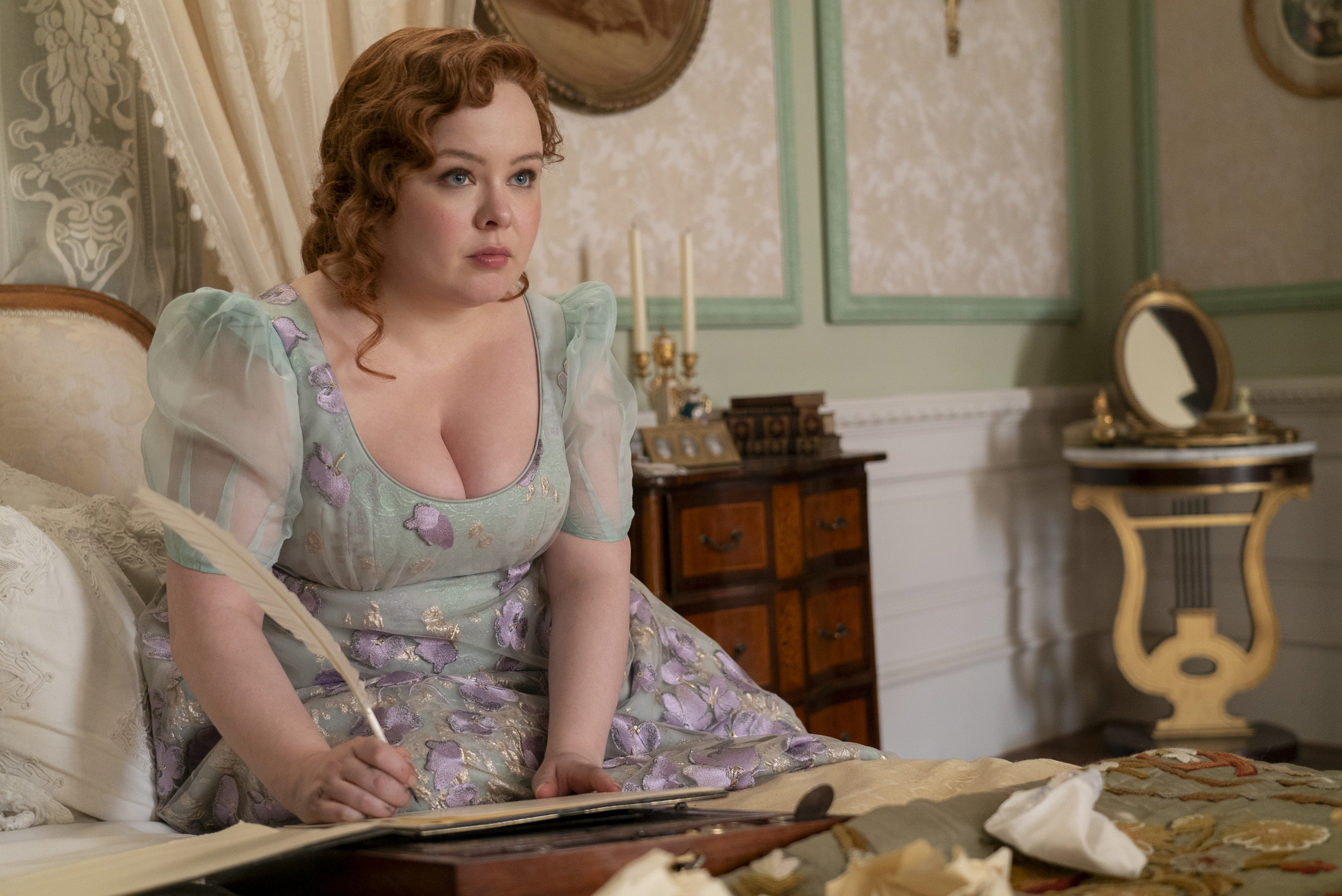 Nicola Coughlan as Penelope Featherington wearing a purple and green dress writing a lady whistledown society paper with a feather pen on a bed in episode 306 of Bridgerton