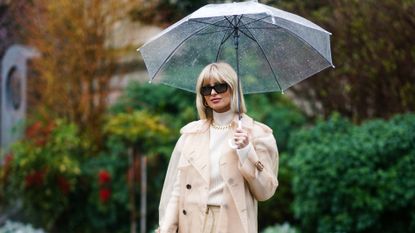 Xenia Adonts wears sunglasses, a white turtleneck pullover, a golden chain necklace, a beige clear long rain jacket, beige pants, outside Maison Margiela, during Paris Fashion Week - Womenswear Fall/Winter 2020/2021, on February 26, 2020 in Paris, France. 