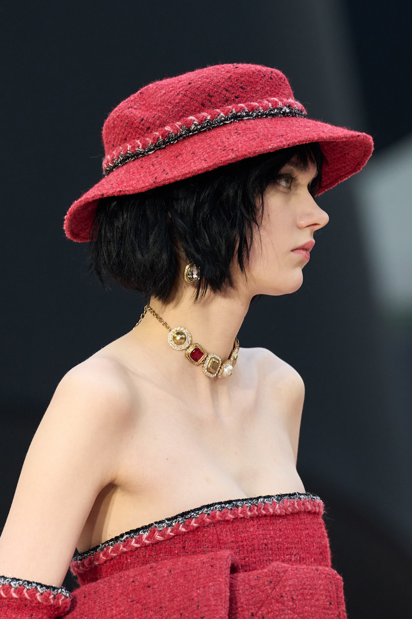 A Chanel model wearing a red tweed bucket hat, a ruby gem gold necklace, and a red tweed dress.