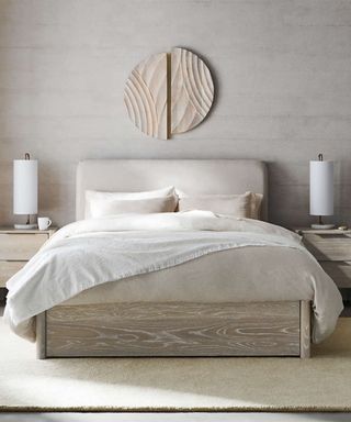 White and beige bedding on a cream bed frame against a beige wall; a white oak nightstand with a white lamp either side.